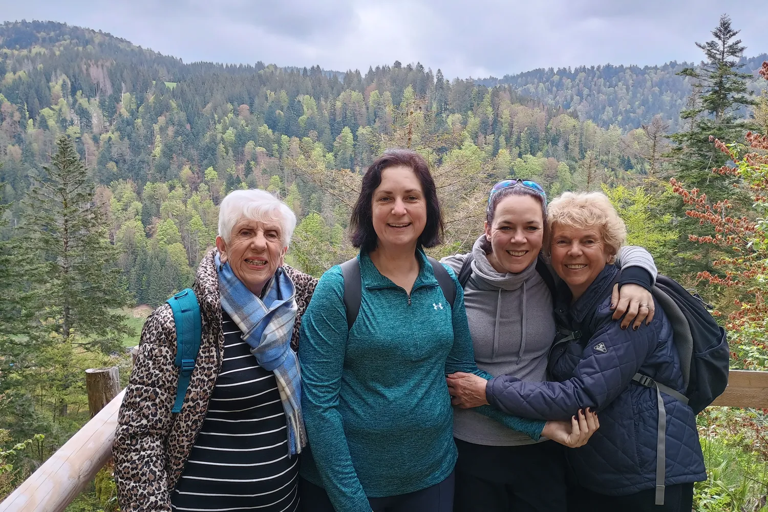 A group of three women and one man smiles together while standing on a wooden bridge surrounded by nature.