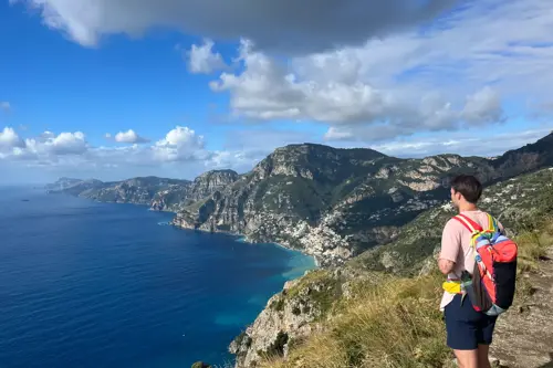 A man with a backpack stands on a scenic trail, gazing out at the vast ocean below him.