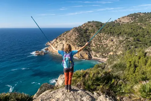 Woman standing on a cliffside rock holding her hiking poles up in the air with back turned to camera