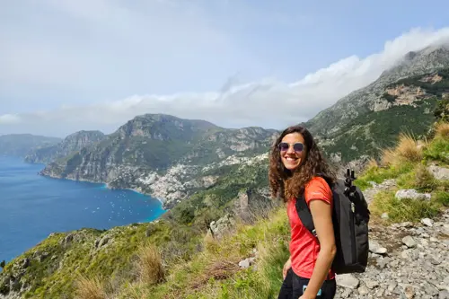 A woman hiking with a backpack pauses on a trail, taking in the stunning ocean view from the Path of Gods.