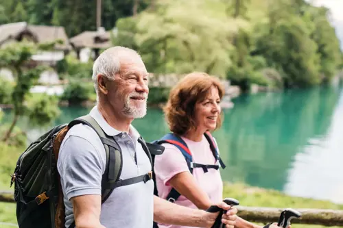 Older man and woman walking with backpacks and hiking poles near a body of water